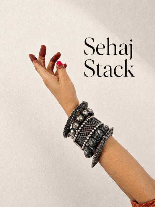 A close-up of a person's raised hand and wrist wearing a stack of thick, textured black and silver beaded bracelets, with the text 'Sehaj Stack' printed on the right against a light beige background.
