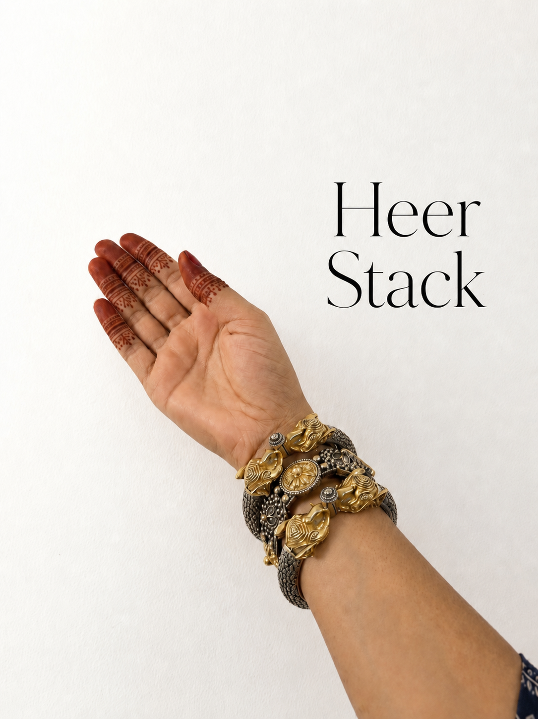 A close-up of a person's hand and wrist wearing a stack of ornate gold and silver-toned statement bracelets, with the text 'Heer Stack' printed on a plain white background.