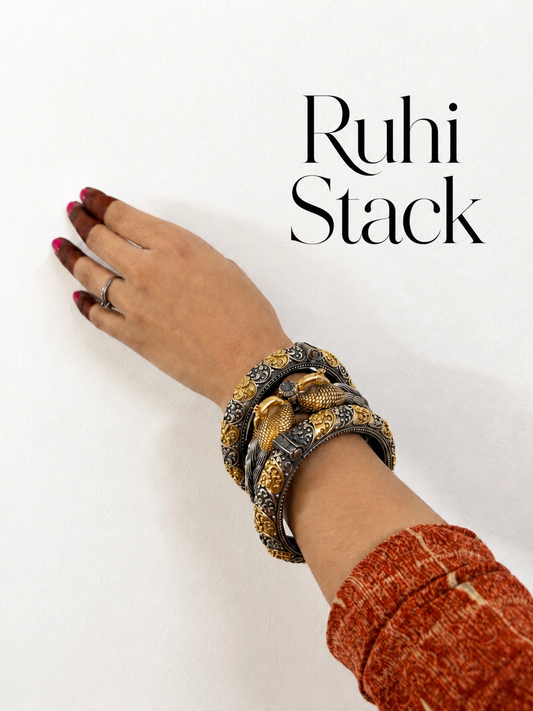 A close-up of a person's hand and wrist wearing a stack of wide, patterned bangles with a gold and dark gray snake design, against a plain white background with the text 'Ruhi Stack' in the upper right.
